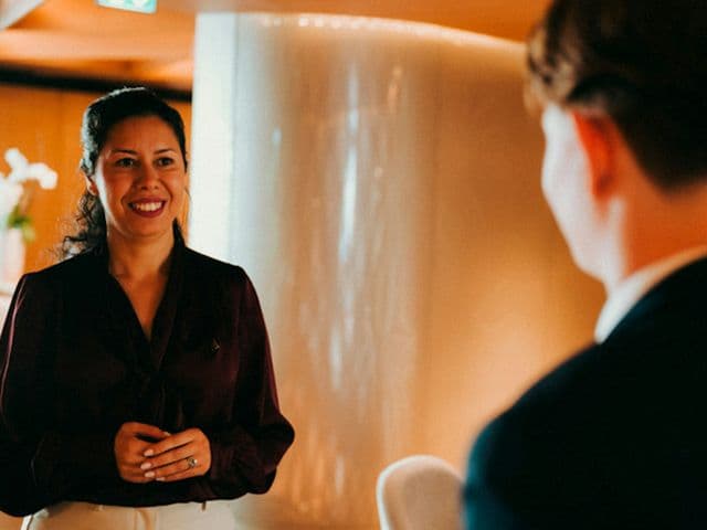 Smiling host in a burgundy blouse greets a suited guest beside a softly lit curved wall at 369 Dine & Bar