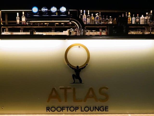 Atlas Rooftop Bar & Loungeの金色のAtlasロゴが入ったバックライト付きバーカウンター、ビールサーバーと酒瓶の棚