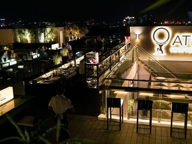 夜のAtlas Rooftop Bar & Loungeの屋上テラス、照明の灯るバーカウンター、ガラスの手すり、スツールと街の夜景