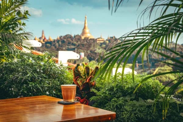 Iced coffee on a wooden table at Café Salween, framed by tropical plants with a golden pagoda on the hill beyond
