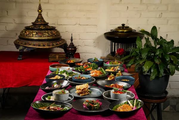 Table spread with many Thai dishes in dark bowls on a magenta cloth, ornate brass vessels behind, at Café Salween