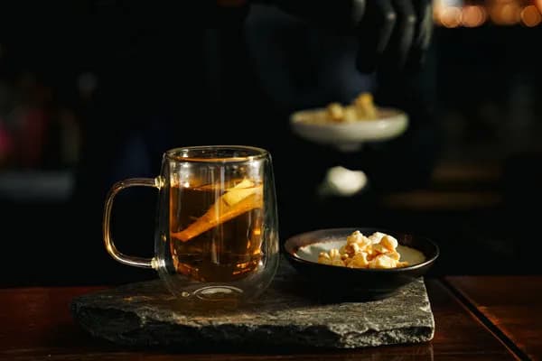 Double-walled glass mug of amber tea with a fruit slice beside a bowl of nuts on a slate board at Café Salween