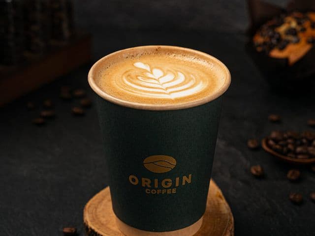 Latte with leaf latte art in a green Origin Coffee paper cup on a wooden coaster, with scattered coffee beans