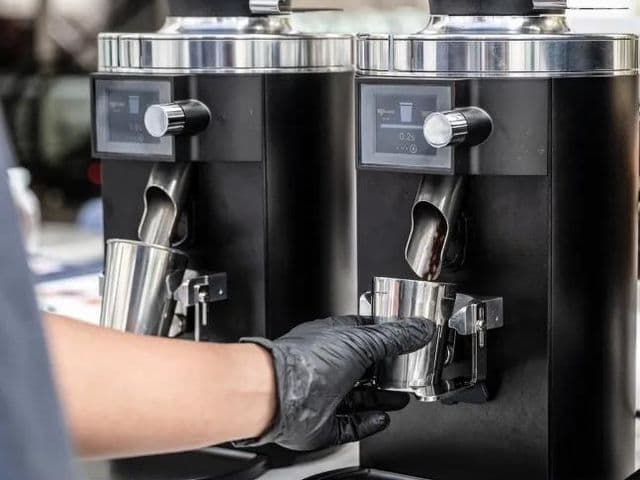 Рука в перчатке держит стальной дозирующий стакан под одной из двух чёрных кофемолок в Pacamara Coffee Roasters
