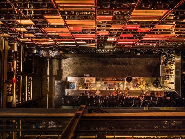 Overhead view of the dimly lit bar counter at Rabbit Hole, with bottles on shelves and a glowing grid ceiling