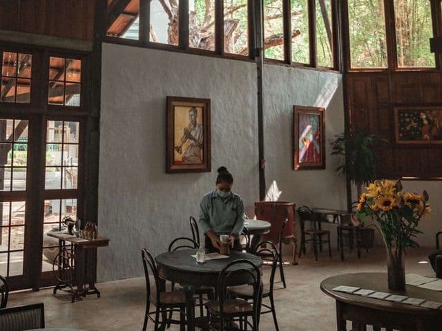 Masked server setting a table in a high-ceilinged café with framed paintings and sunflowers at Rangoon Tea House