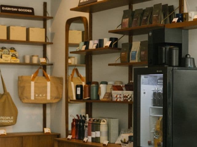 Rootsの物販コーナー、コーヒー豆の袋やタンブラー、トートバッグが並ぶ木製棚と明るいドリンク冷蔵庫
