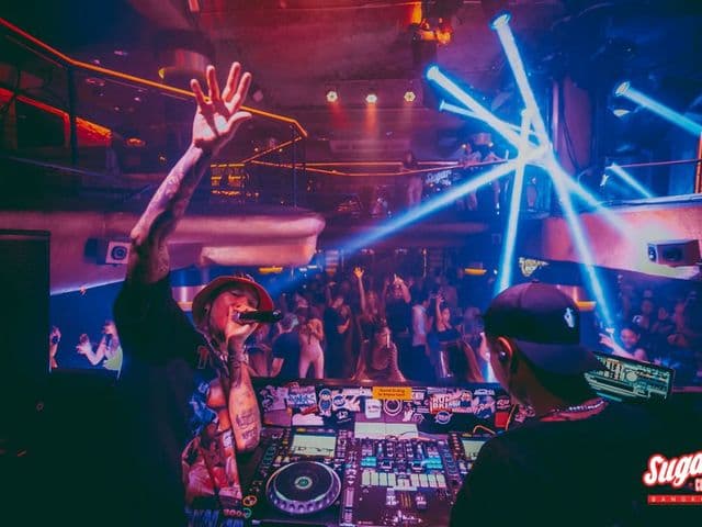 Sugar Club Bangkokで、青いレーザーの下で踊る観客に向けDJブースで手を挙げるタトゥーのMC