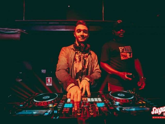 赤い照明の下、Sugar Club Bangkokでストライプのジャケットを着たDJがプレイし、後ろに男性が立っている