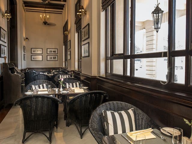 The Strand Café の高い窓辺に並ぶ、ストライプのクッション付き黒い籐椅子とセッティング済みのテーブル