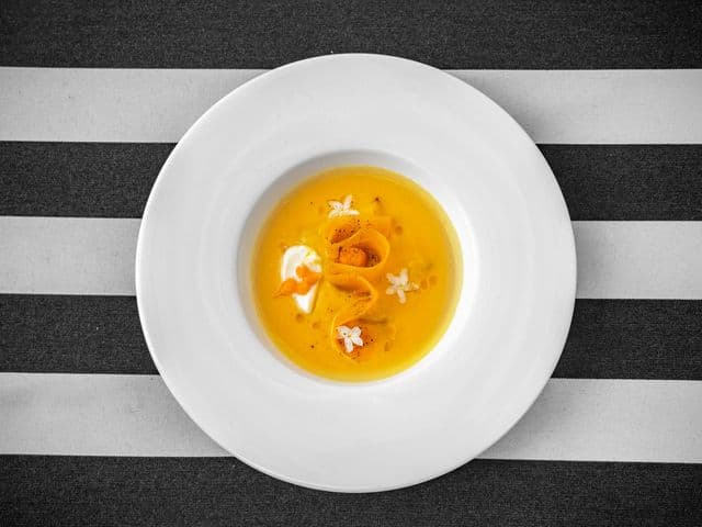 The Strand Café の縁広の白い皿に盛られた、リボン状のにんじん、クリーム、白い小花を添えた黄金色のスープ