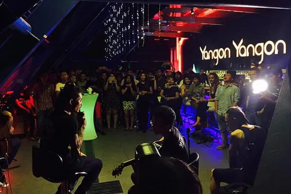 Crowd watching an acoustic guitar performance under red and blue lights at Yangon Yangon rooftop bar