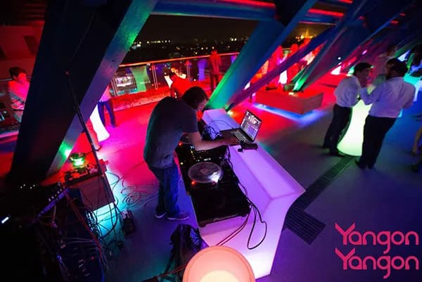 DJ at turntables and laptop on a neon-lit rooftop terrace at Yangon Yangon, with guests and a city night view