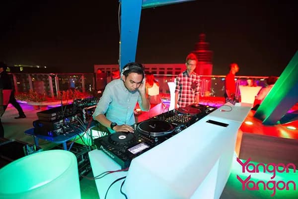 DJ in headphones spinning at a glowing white booth on the Yangon Yangon rooftop at night, with guests behind