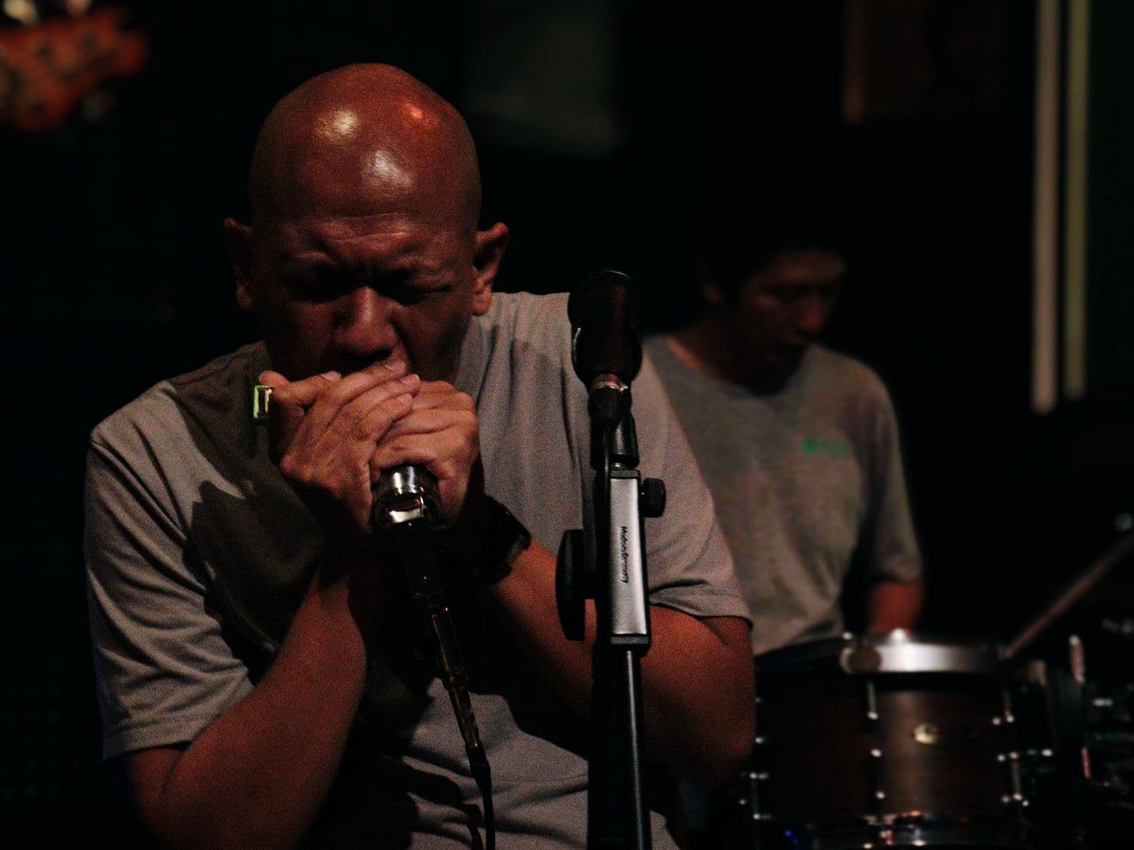 Bald musician playing harmonica into a microphone with a drummer behind him at Bangkok Mojo