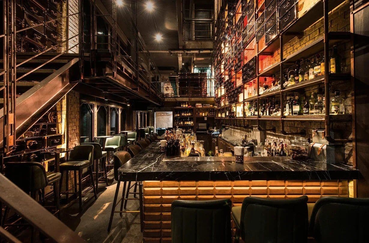Dimly lit bar at Rabbit Hole with a black marble counter, green stools and tall shelves of backlit bottles