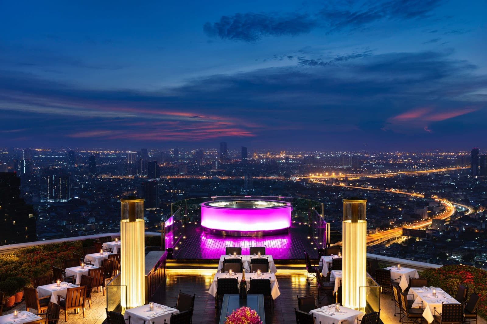 Rooftop terrace at Sky Bar with white-clothed tables, a glowing purple bar and a city skyline at dusk